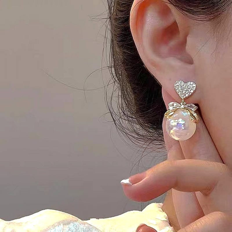 dazzle21 gold heart crystal stud earrings with a delicate bow and lustrous faux pearl drop worn on an ear